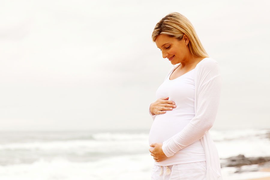 Mulher grávida de pé olhando sua barriga, com as mãos pousadas sobre a mesma, à beira mar